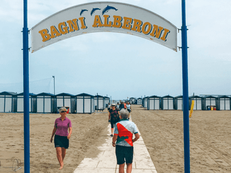 Folk går under en bue med teksten 'Bagni Alberoni' mod en sandstrand med rækker af blå og hvide strandkabiner under en overskyet himmel.