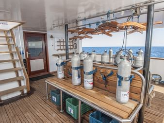 Dykkedæk på MV Tala med iltflasker, træudstyrsbord, trappe til øvre dæk og havudsigt med klart blåt vand.