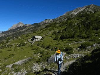 Vandrer med orange hat og rygsæk på bjergsti i Queyras, Frankrig. Grønne alpenge, bjerghy og klippefyldte toppe under blå himmel.