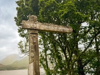 Træskilt for West Highland Way ved en skotsk sø, med grønne træer og tågede bakker i baggrunden.