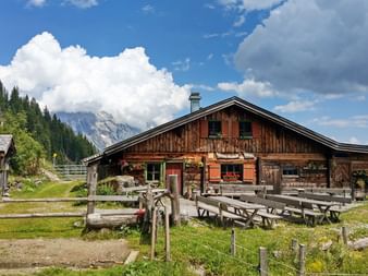 Wooden alpine hut with outdoor seating area in Austrian Alps. Mountain peaks and forest visible in background under blue sky with white clouds.
