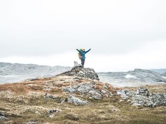 Vandrer med løftede arme på stenvarde på Hardangervidda-plateau. Sparsom vegetation og snedækkede bjerge i baggrunden.