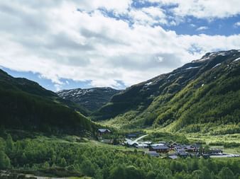 Liten fjellbosetting Tyristhytte i grønne Aurlandsdalen, omgitt av skogkledde skråninger og snødekte fjell under skyet himmel.