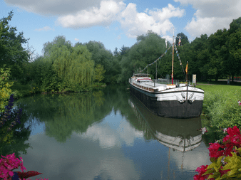 Sort historisk pram fortøjet på rolig kanal i Strasbourg, omgivet af grønne træer og farverige blomster langs vandkanten.