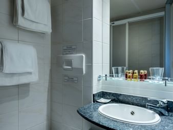 Moderne badeværelse på MS Olympia med hvid oval vask i mørk marmorplade, spejl, toiletartikler og hvide håndklæder på flisevægge.