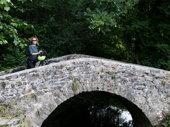 Cyclist with helmet and bicycle resting on historic stone arch bridge surrounded by lush green forest vegetation.