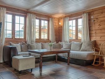 Træhytte interiør med hjørnesofa, sofabord og tre vinduer med hvide gardiner. Stengulv og træpaneler skaber varm atmosfære.