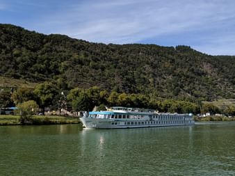 Hvidt flodkrydstogtskib Switzerland sejlende på roligt grønt vand med skovklædte bakker og lille landsby i baggrunden under blå himmel.