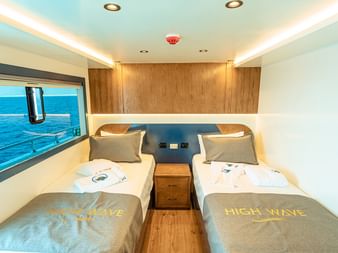 Moderne dobbeltkahyt ombord på High Wave med to enkeltsenge, vindue med havudsigt, træpaneler og blåt hav udenfor.