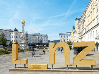 Store gyldne bogstaver LINZ på en brostensplads med historiske bygninger, barokkesøjle, cyklister og fodgængere under blå himmel.
