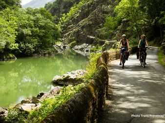 Two cyclists riding along a stone-walled path beside a green river, surrounded by lush vegetation and rocky cliffs in the Bordeaux region.