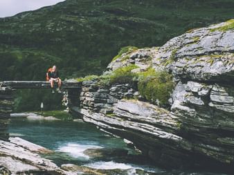 Fotturist sittende på steinbro over turkis elv i Aurlandsdalen, Norge. Lagdelte fjellformasjoner og grønne fjell i bakgrunnen.