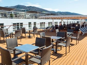 Træ soldæk på flodkrydstogtskib med kurvemøbler, borde og liggestole. Bjerge og landskab synlige i baggrunden.