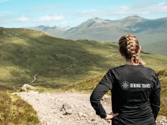 Kvinde med flettet hår i Bering Travel jakke står på sti med udsigt over grønne skotske højlande med bjerge under blå himmel.