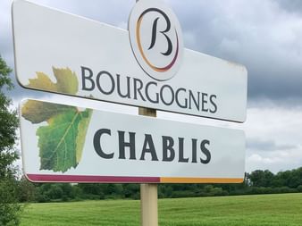 White road sign displaying 'BOURGOGNES CHABLIS' with grape leaf logo and colorful border, set against green countryside landscape.
