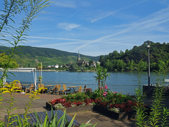 Flodterrasse i Boppard med gule stole, farverige blomsterbede og udsigt over Rhinen med vingårde på skråningen og landsby.