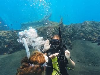 To dykkere under vann nær et korallrev på Bali. En dykker vinker til kameraet mens luftbobler stiger opp i det klare blå vannet.
