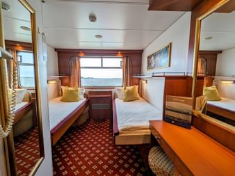 Interiør af to-sengs kahyt på MS Rigoletto med to enkeltsenge, vindue med gardiner, træskrivebord, stol og rødt tæppe.