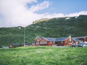 Rød DNT-turisthytte i Aurlandsdalen med grønne bjerge i baggrunden. Køretøjer parkeret ved siden af hovedbygningen.