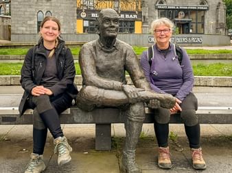 To kvindelige vandrere sidder på en bænk ved siden af en bronzestatue i en skotsk by. Stenbygninger og en pub er synlige i baggrunden.