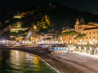 Natbillede af en Amalfikysten by med oplyste bygninger langs vandkanten, en kirke med klokketårn og lys reflekteret i havet.