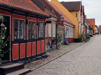 Kullerstensgata kantad av traditionella korsvirkeshus i rött, gult och vitt på Ærø, Danmark. Cyklar och blommor pryder fasaderna.