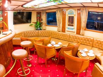 Indvendig lounge på Donau-krydstogtskib med orange lædersæder, runde borde, barområde og rødt tæppe. Vinduer viser aftensudsigt.