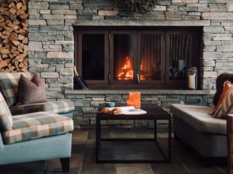 Moderne hotelllounge med steinpeis, brennende ild, stablet ved, rutete lenestol, lærsofa og sofabord med stearinlys.