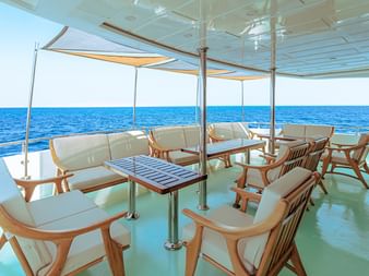 Overdækket dæk på High Wave liveaboard skib med træstole, borde og hvide puder med udsigt over blåt hav under klar himmel.