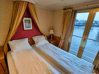 Kahyt med to senge på MS Casanova med hvidt sengetøj, gyldne gardiner, rød accentvæg og store vinduer med udsigt over havnen.