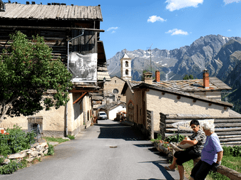 To vandrere hviler i en traditionel alpelandsby med stenbygninger, et kirketårn og dramatiske bjergtinder i baggrunden.