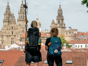 Two hikers with backpacks viewing Santiago de Compostela Cathedral from a rooftop. The cathedral's towers rise above red-tiled roofs.