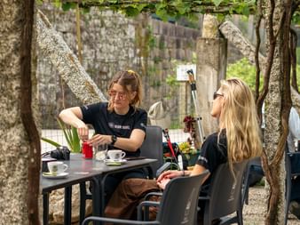 Two women in black Bering Travel shirts sitting at outdoor café table under trees. Stone wall visible in background. Portuguese Camino day 12.