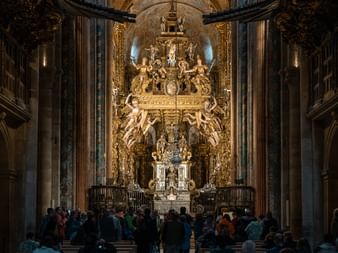 Utsmykket barokk katedralinteriør med gyllen altertavle, steinsøyler og pilgrimer samlet i skipet på Camino de Santiago.