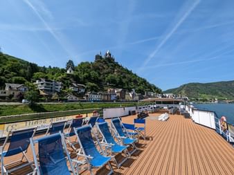 Soldæk på MS Olympia med blå liggestole på trægulv, med udsigt over Rhindalen med slotskronet bakke og flodby.