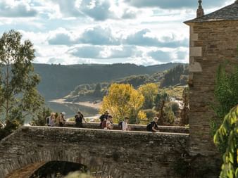 Gruppe Camino-pilgrimer hviler på gammel steinbro med buer. Bølgende åser og høsttrær synlige i bakgrunnen under skyet himmel.