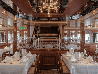 Elegant skibsrestaurant med hvide duge, træpaneler og central trappe. Reflekterende loft og panoramavinduer langs væggene.
