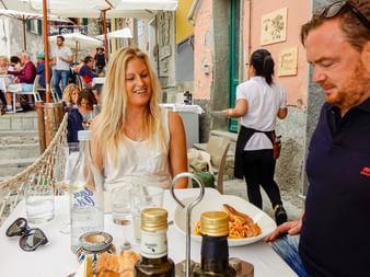 Blonde woman in white top smiling at outdoor restaurant table while waiter in dark shirt serves pasta. Colorful Italian village buildings visible.