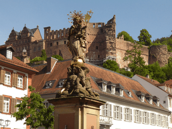 Gylden statue på stensokkel på Heidelbergs torv med historiske slotsruiner på bakken over traditionelle bygninger.