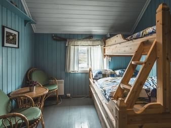 Koselig soverom i Geiterygghytta DNT-hytte med køyesenger i tre, blå panelvegger, hvitt tak og kurvstolene ved vinduet.