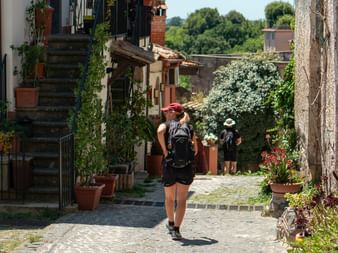 Two hikers with backpacks walking through a cobblestone alley in an Italian village. Stone houses with flower pots line the narrow street.