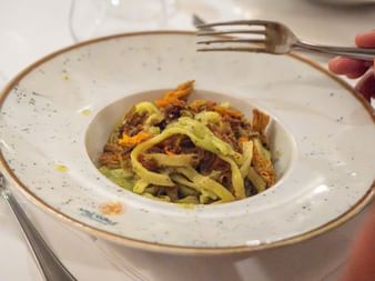Hand holding fork over plate of fresh pasta with meat sauce served on white speckled plate with gold rim in Italian restaurant.
