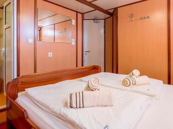 Skibskahyt med dobbeltseng, hvidt sengetøj, foldede håndklæder, træpaneler og eget badeværelse med vask synligt gennem åben dør.