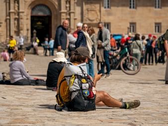 Pilgrim with backpack sitting on stone plaza in Santiago de Compostela. Historic church entrance and crowds of people visible in background.