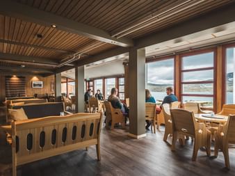 Interiør i DNT Finsehytta hytte med træmøbler, ribbeloft og store vinduer mod bjergsø. Gæster sidder ved borde.