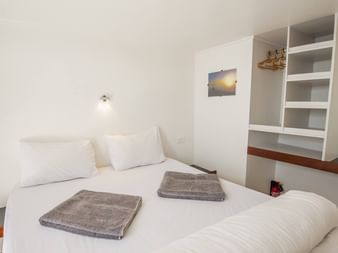 Ren hvid skibskahyt med dobbeltseng, hvidt sengetøj, grå håndklæder, indbyggede hylder og indrammet kunst på væggene.