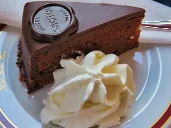 Trekantet stykke chokolade Sachertorte med mørk chokoladeglasur og Hotel Sacher segl, serveret med dekorativ flødeskum på hvid tallerken.