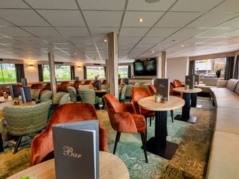 Interiør af MS Princess lounge med orange fløjlslænestole, grønne mønstrede stole, runde borde og væg-TV. Store vinduer viser udendørs udsigt.