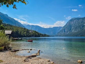 Stenstrand med klart turkost vatten omgivet av skogklädda berg i Juliska alperna, Slovenien. En röd båt flyter nära ett båthus.