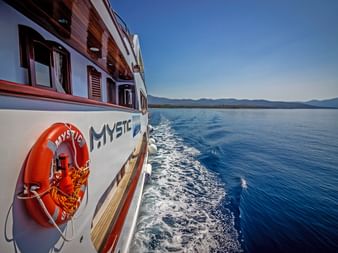 Sidovy av fartyget MY Mystic med orange livboj seglande genom blått Adriatiskt vatten. Kroatisk kustlinje med berg synlig i bakgrunden.